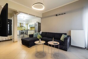 A modern living room with a black sofa, two small round tables, a TV, and a kitchen in the background. - Faliro Escape - Spacious Stay in Thessaloniki (Thessaloniki)