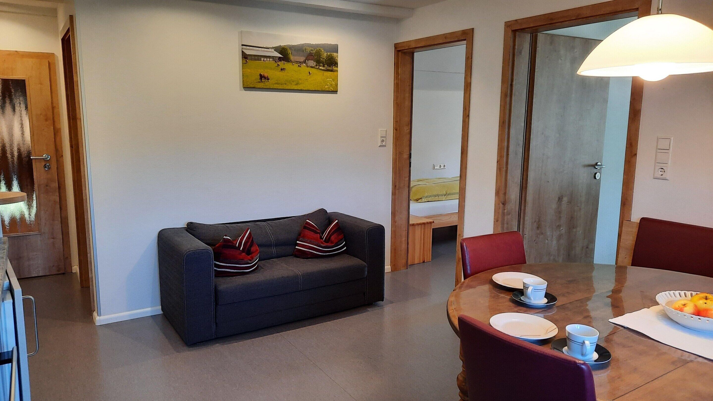 A room with a sofa, a dining table with chairs, a painting on the wall, and a door leading to another room.