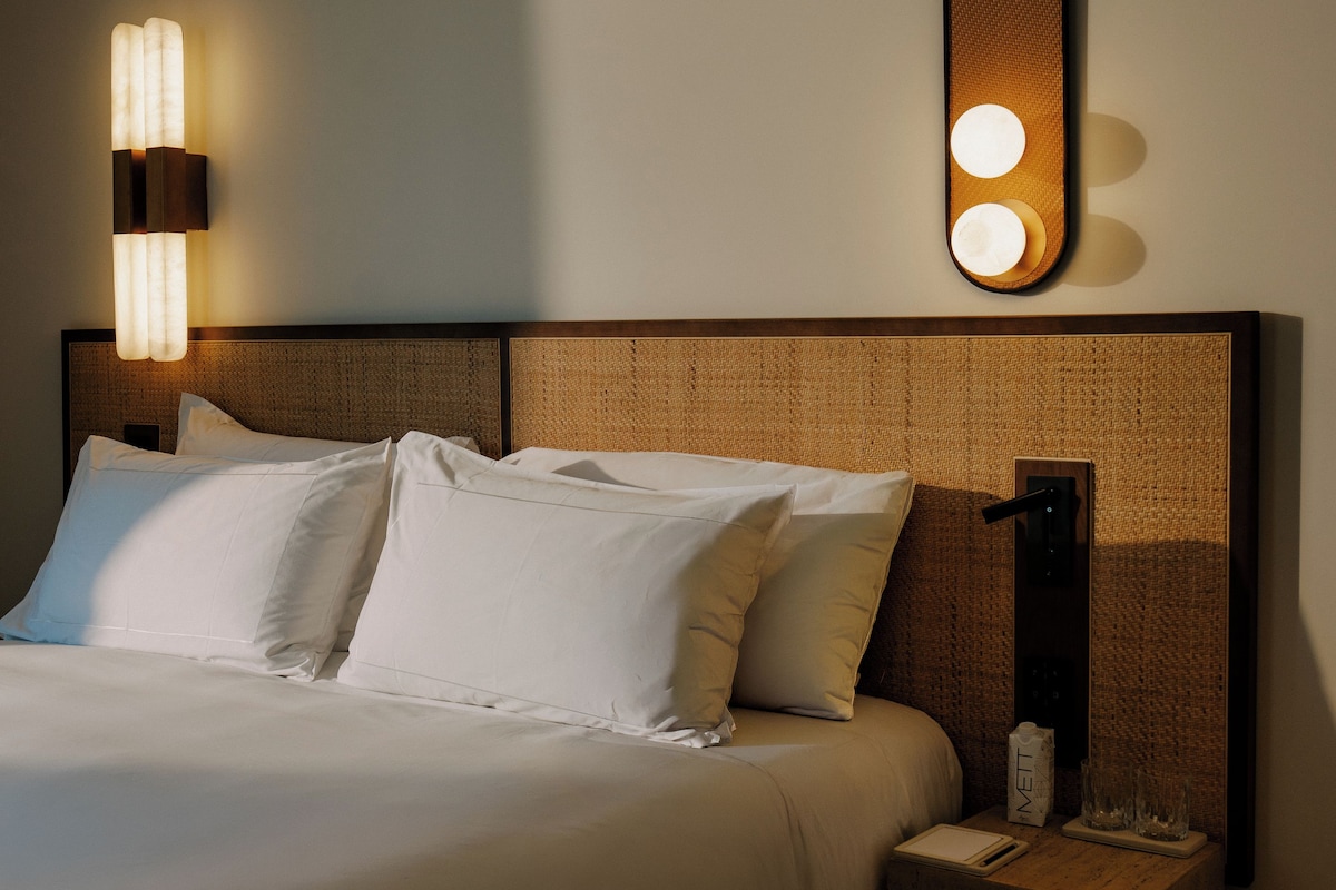 Una cama bien tendida con sábanas blancas, un cabecero con diseño texturizado, una mesita de noche con lámpara y una lámpara de pared.