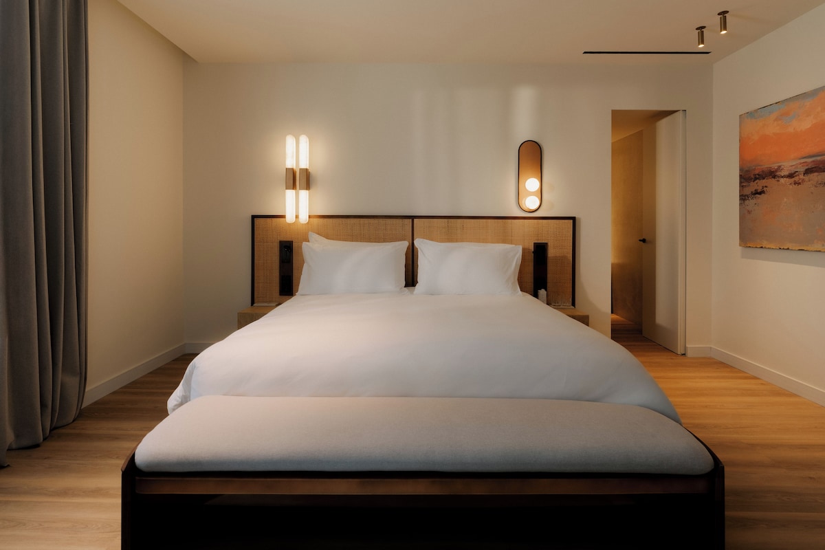 A modern hotel room with a large bed, wooden headboard, two bedside lamps, and a painting on the wall.