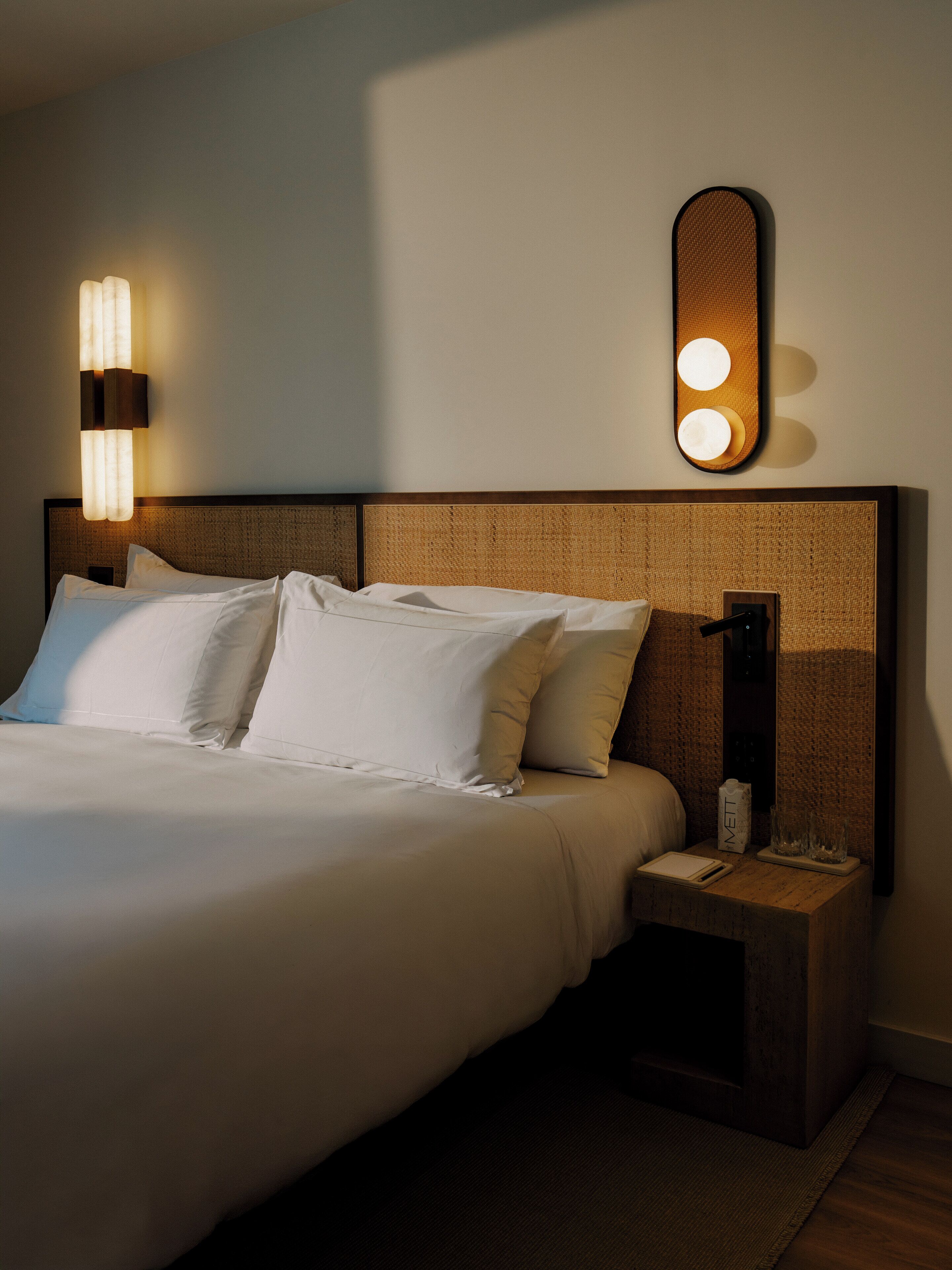 Una cama bien tendida con sábanas blancas, un cabecero con diseño texturizado, una mesita de noche con lámpara y una lámpara de pared.