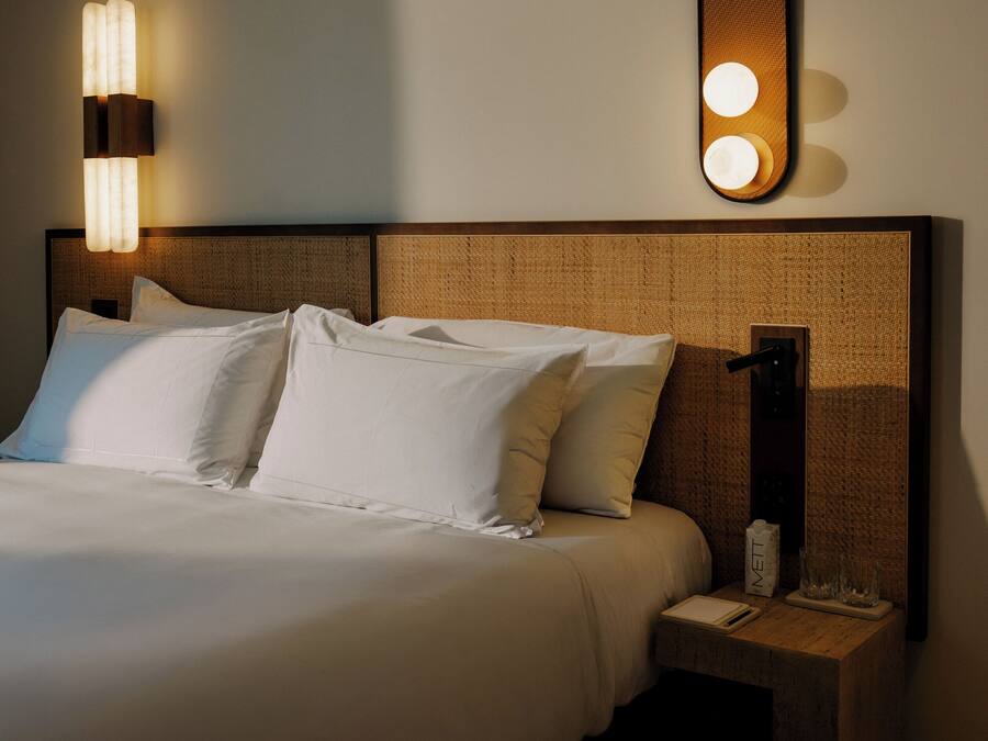 A neatly made bed with white linens, a headboard with a textured pattern, a bedside table with a lamp, and a wall-mounted light fixture.