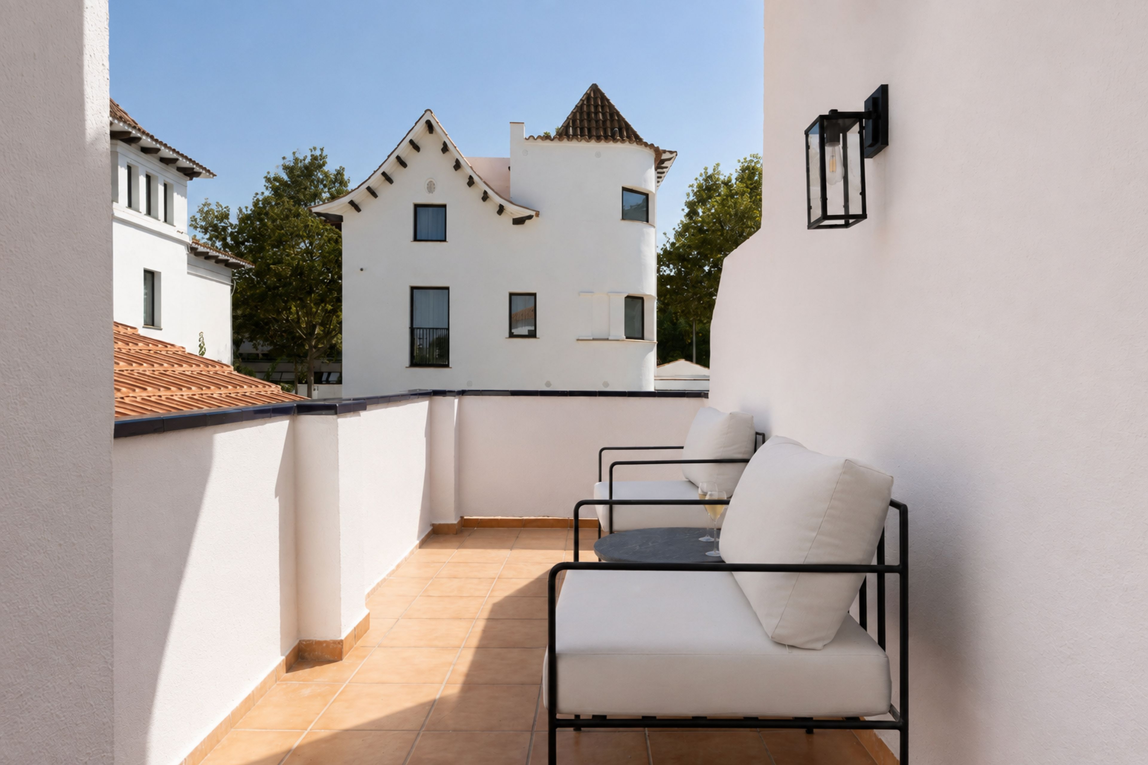 A balcony with two white sofas and a table, a tiled floor, and a white building with a tiled roof in the background.