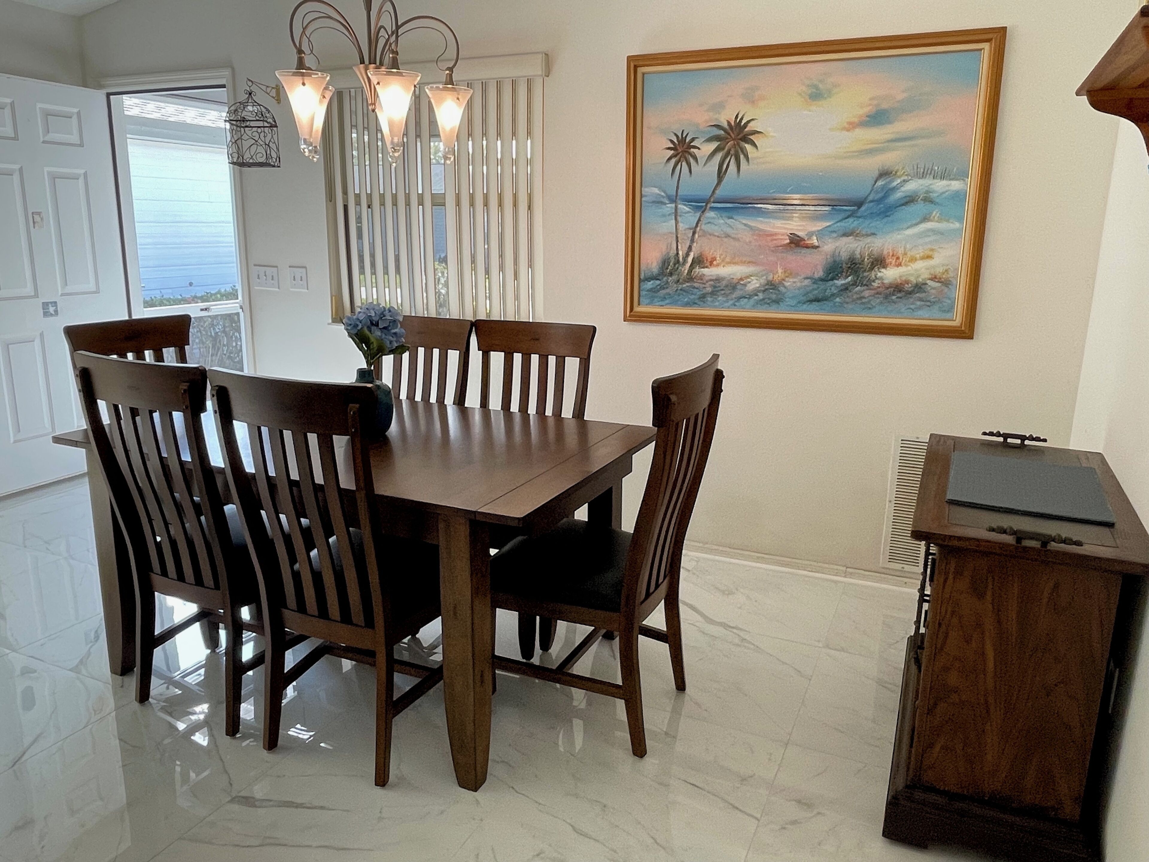 A dining area with a wooden table and chairs, a vase with flowers, a painting of a beach scene, and a small wooden cabinet.