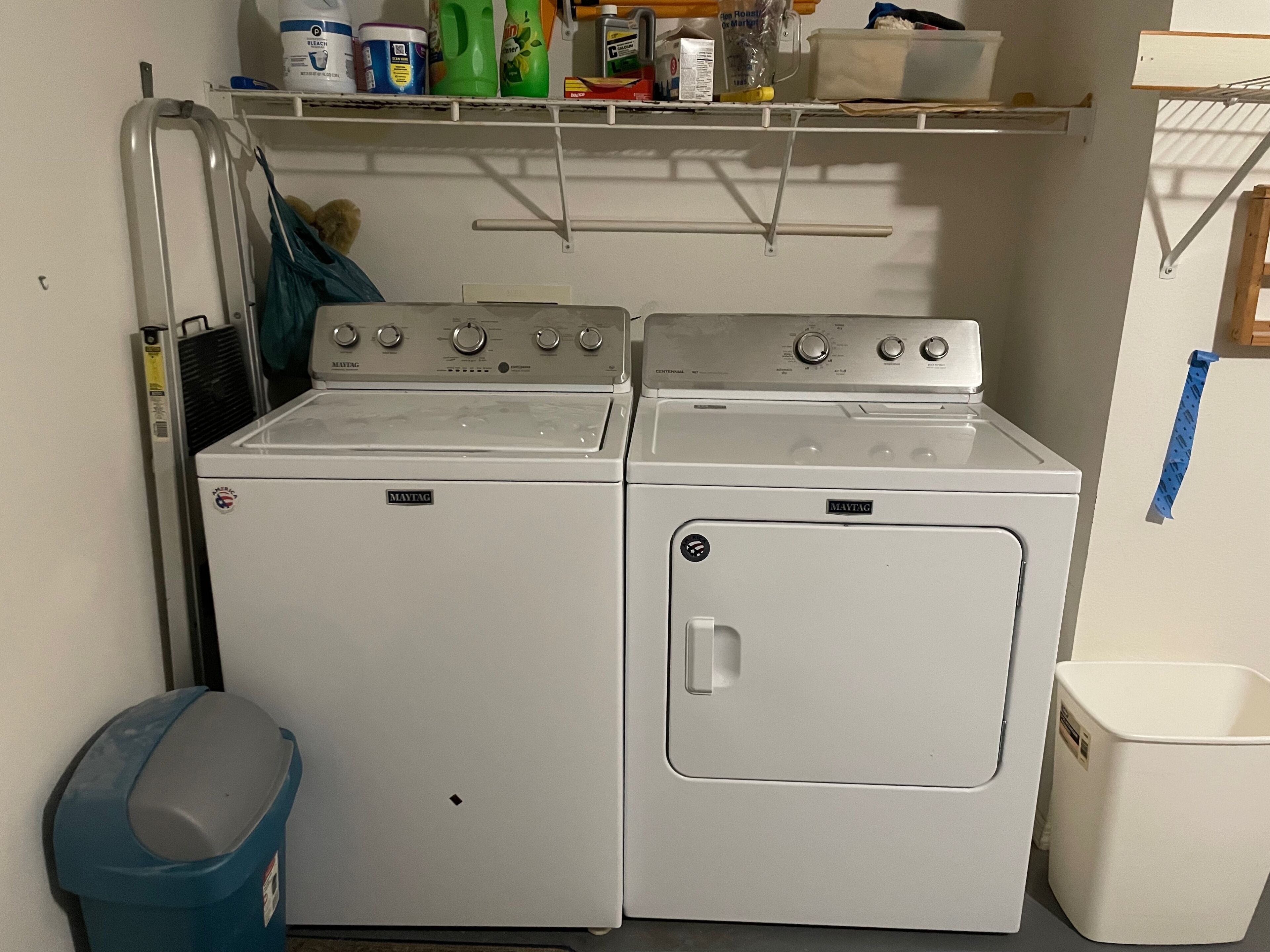 Un área de lavandería con dos lavadoras y secadoras Maytag, un basurero azul y un estante con útiles de limpieza.