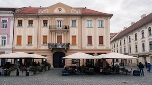 Un bâtiment historique doté d’une entrée centrale, d’un balcon et d’un espace extérieur aménagé avec des parasols.