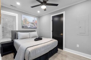 Une chambre à coucher comprenant un lit, un ventilateur de plafond, une fenêtre avec des stores, une porte et un interrupteur fixé au mur.