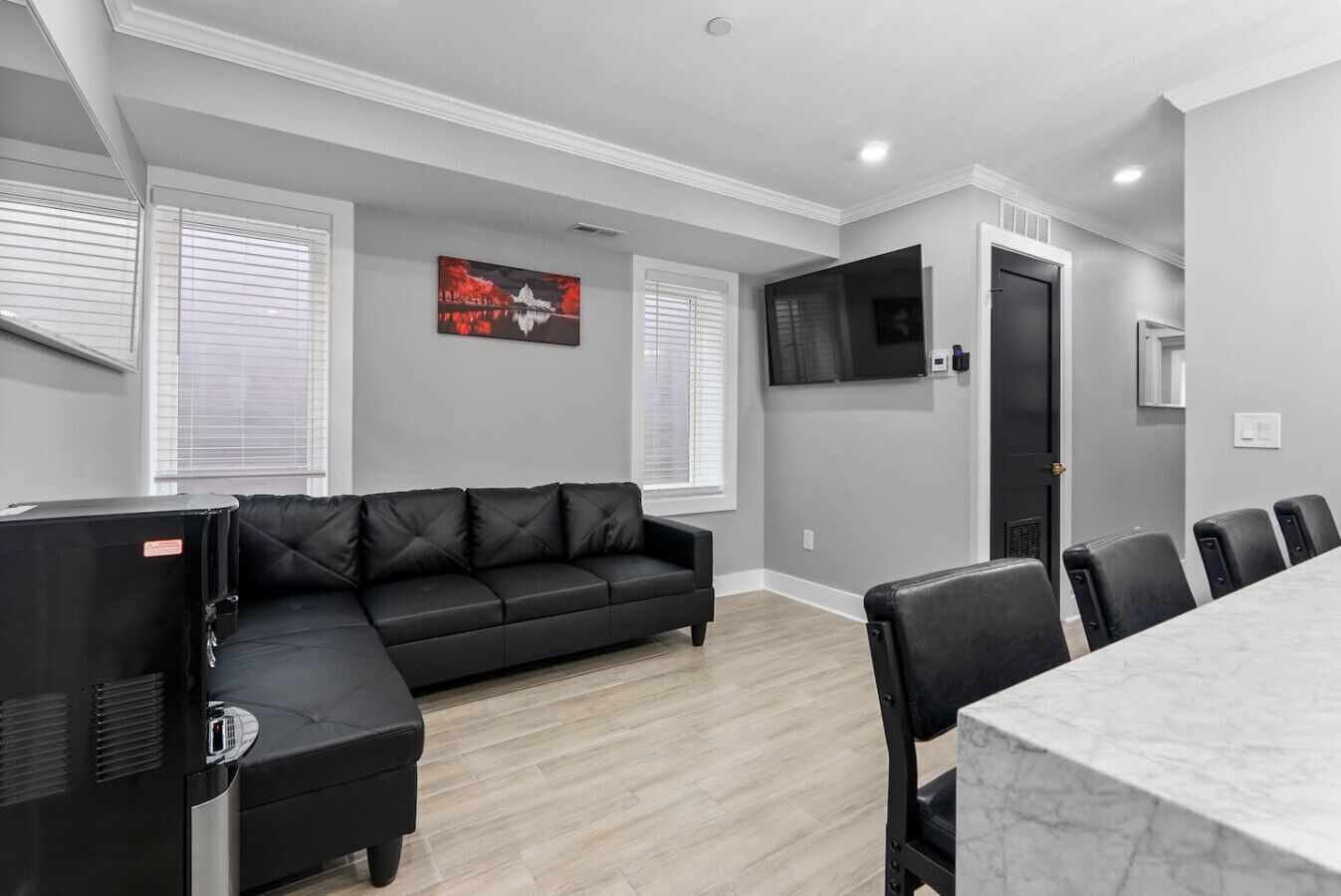 A modern living room with a black leather sofa, a TV mounted on the wall, and a dining area with a marble table and chairs.