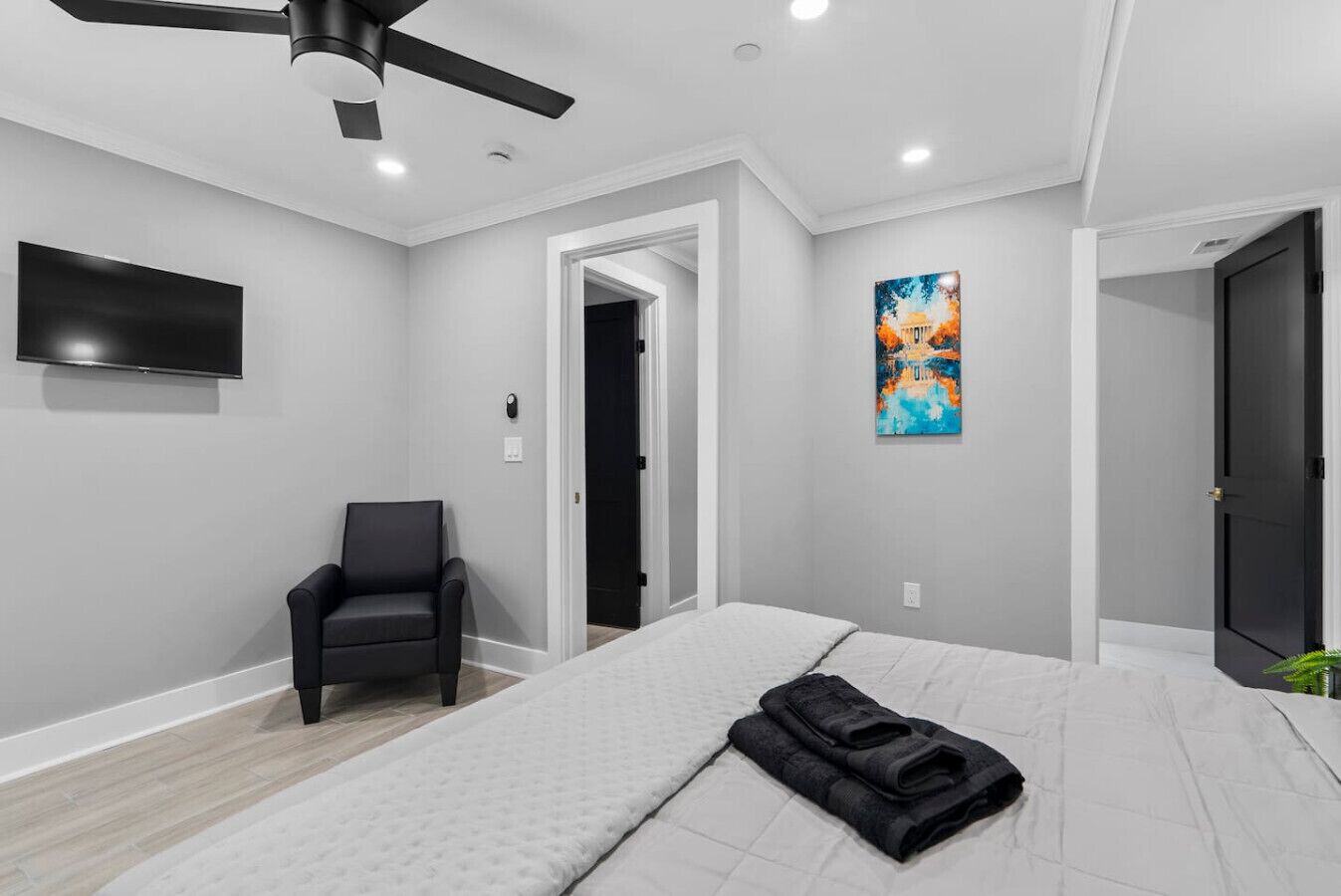 Un dormitorio moderno con cama, sillón, ventilador de techo, televisor y un cuadro.