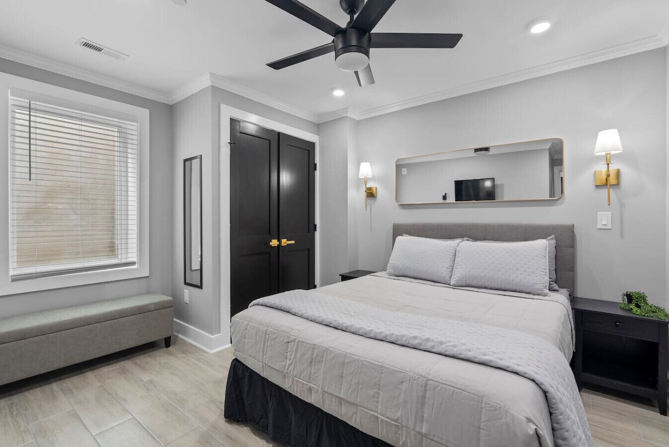 A bedroom with a bed, a ceiling fan, a bench, a door, and a wall-mounted TV.