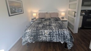 Quarto com cama, duas mesas de cabeceira com luminárias, um quadro na parede e uma porta de correr para uma cozinha.