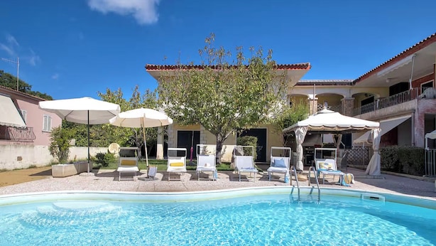 Una piscina con lettini e ombrelloni, circondata da edifici con tetto in tegole.