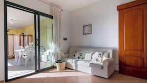 Una stanza con una porta scorrevole in vetro che dà su un'area esterna con tavolo e sedie, un divano bianco con cuscini decorati, una porta in legno e un quadro appeso al muro.