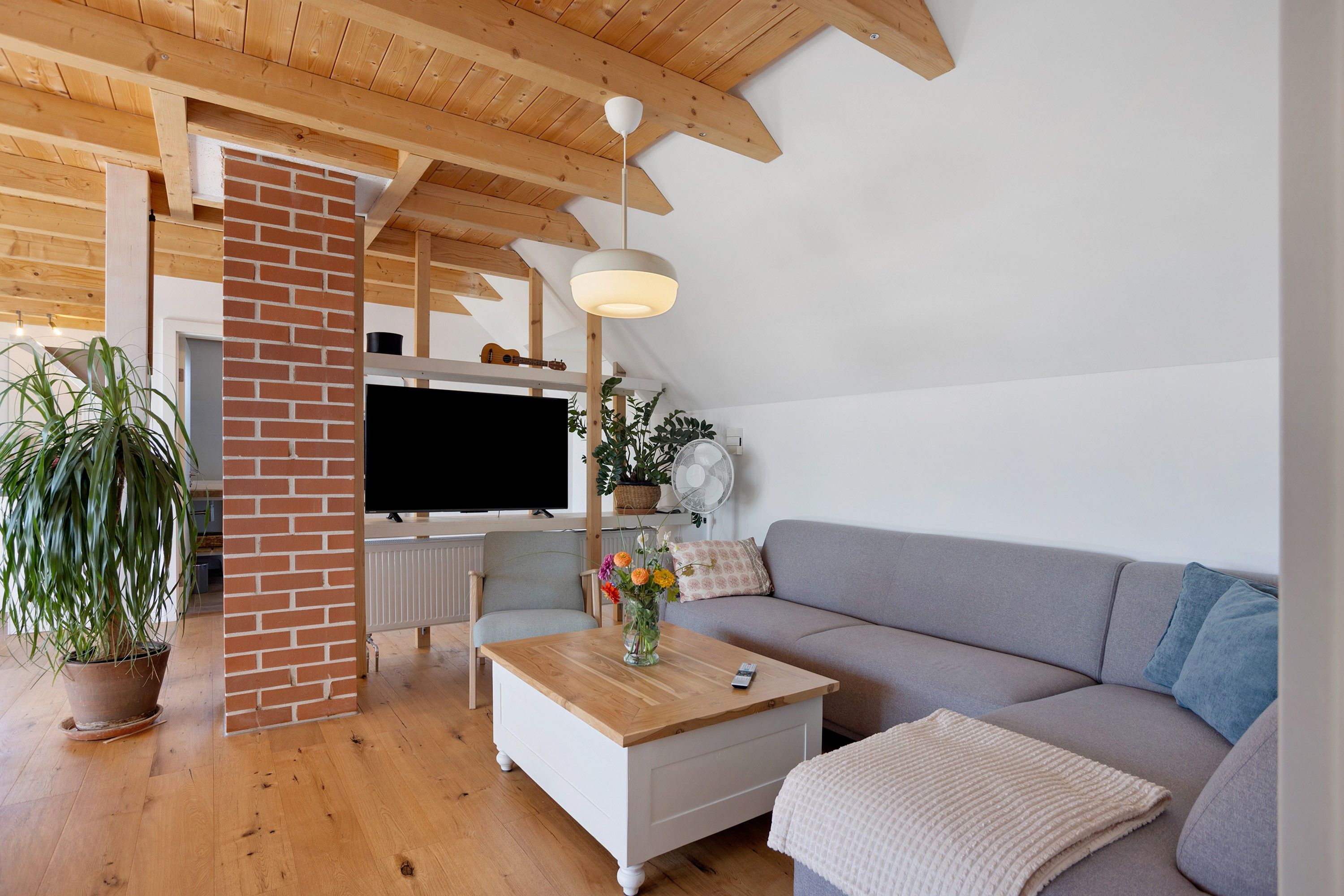 A modern living room with a wooden ceiling, a brick wall, a flat-screen TV, a sofa, a coffee table, and a potted plant.