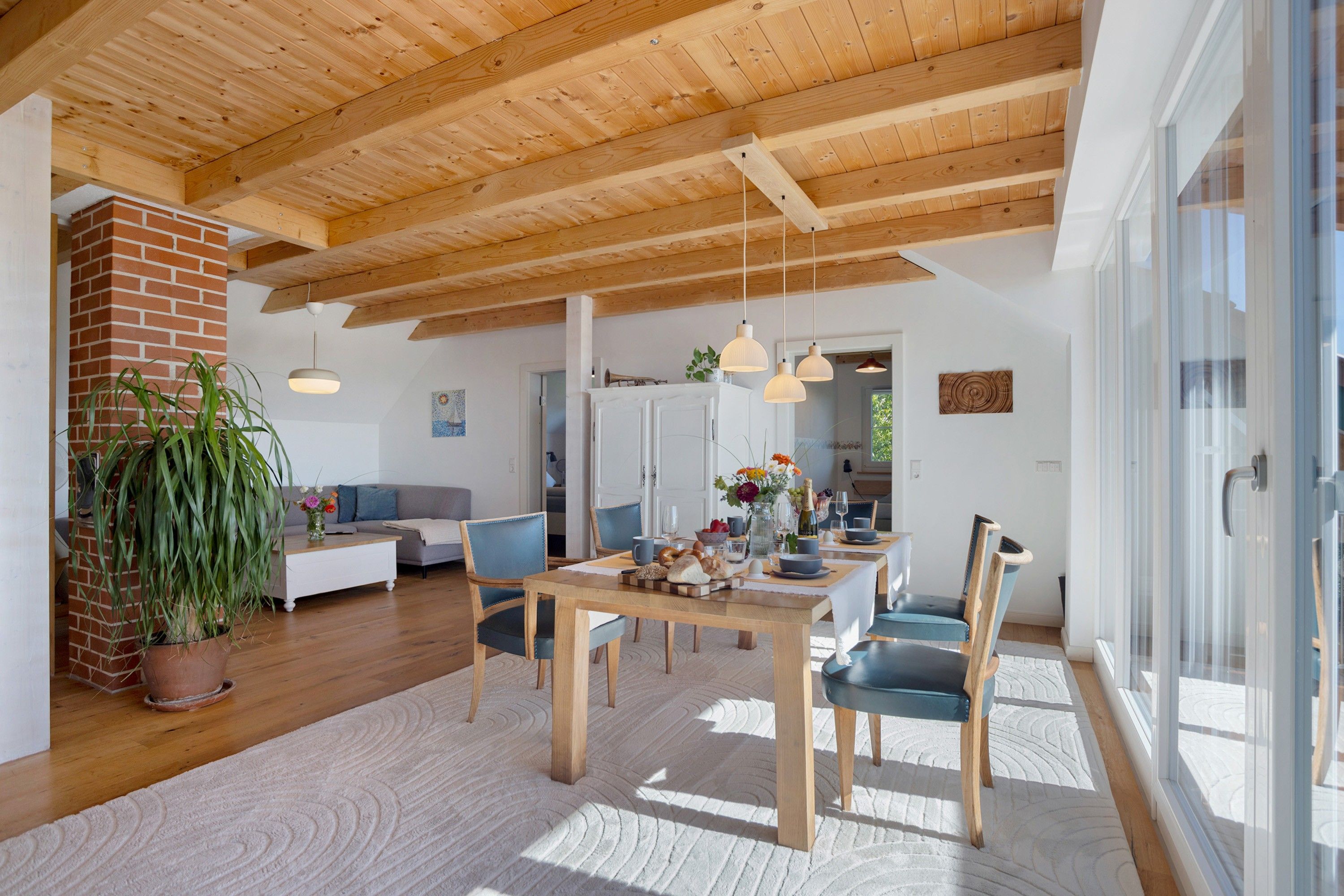 A spacious living area with a wooden ceiling, a dining table set for four, and a large window providing natural light.