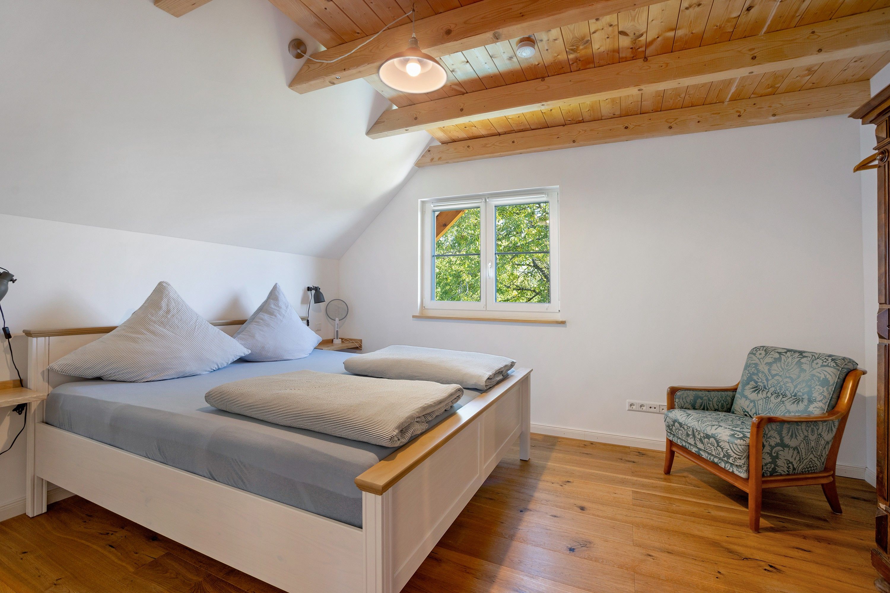 A bedroom with a wooden ceiling, a bed with pillows, a chair, and a window with a view of trees.