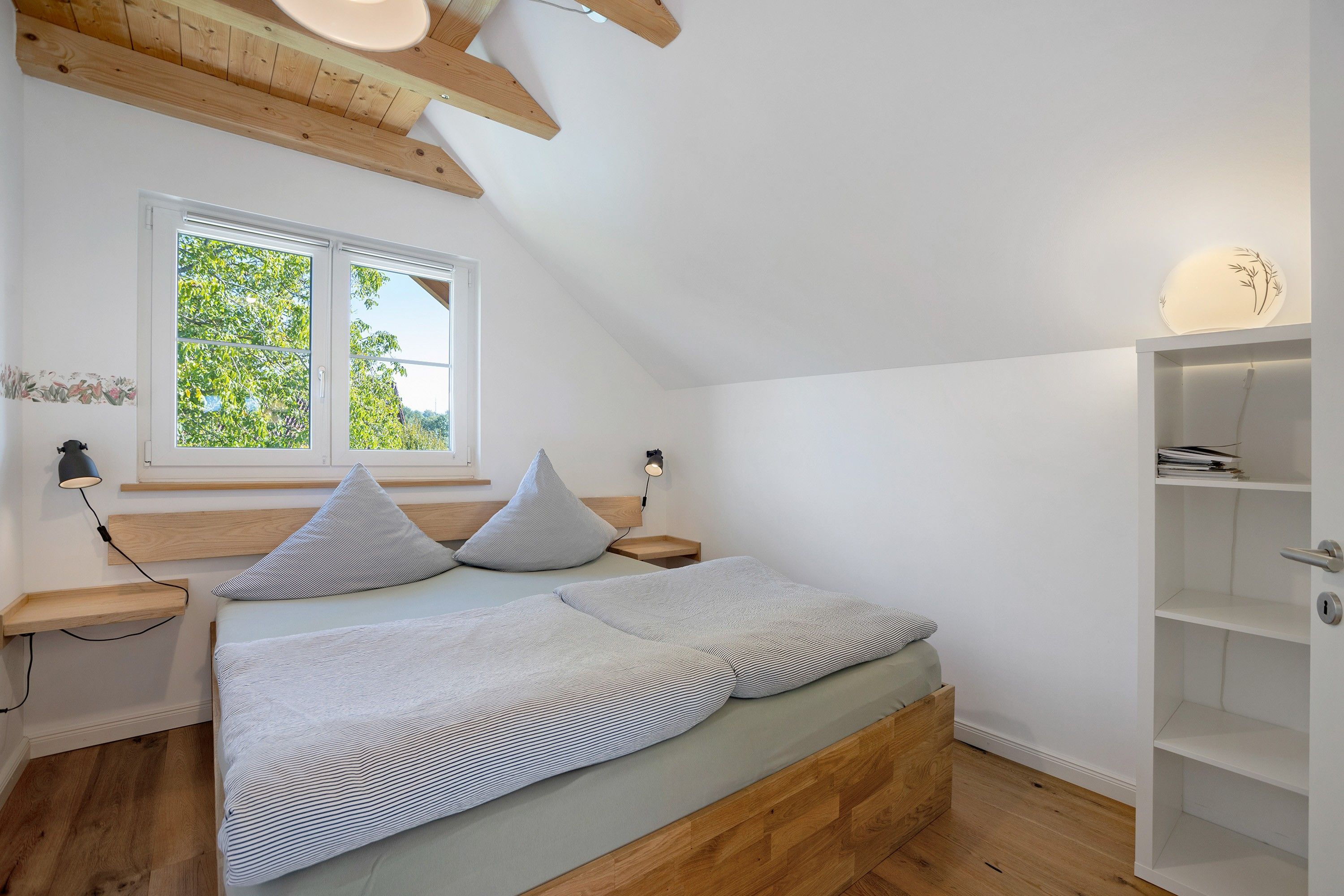 Een slaapkamer met een schuine kap, een bed met twee kussens, een nachtkastje, een raam met uitzicht op groen, en een wandplank.