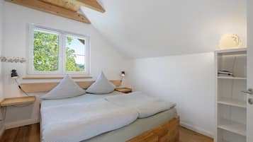 Een slaapkamer met een schuine kap, een bed met twee kussens, een nachtkastje, een raam met uitzicht op groen, en een wandplank.