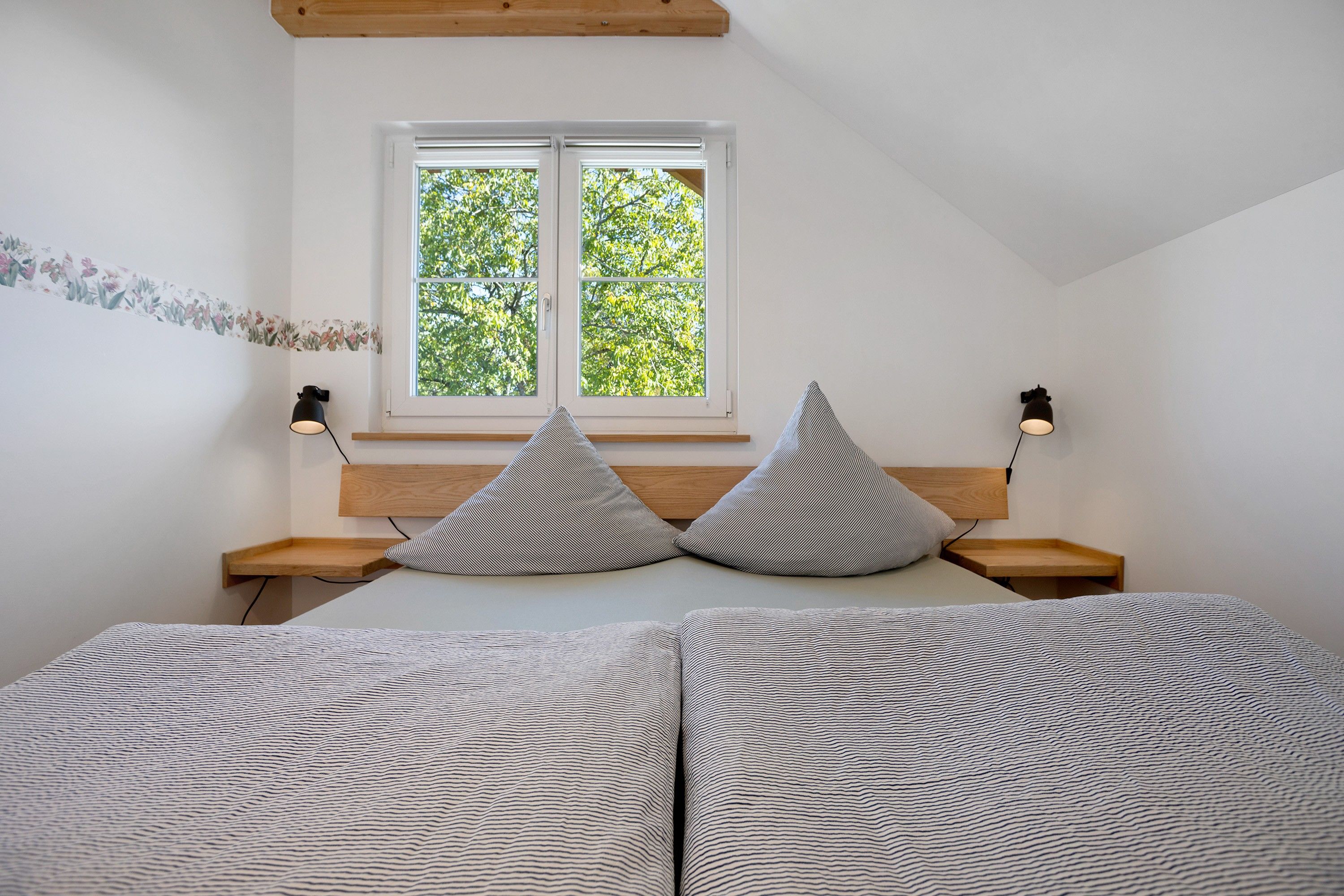 A bedroom with a bed, two pillows, a nightstand, a window, and a wall-mounted lamp.