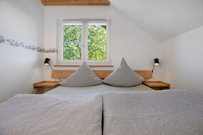 Un dormitorio con cama, dos almohadas, una mesita de noche, una ventana y una lámpara de pared.
