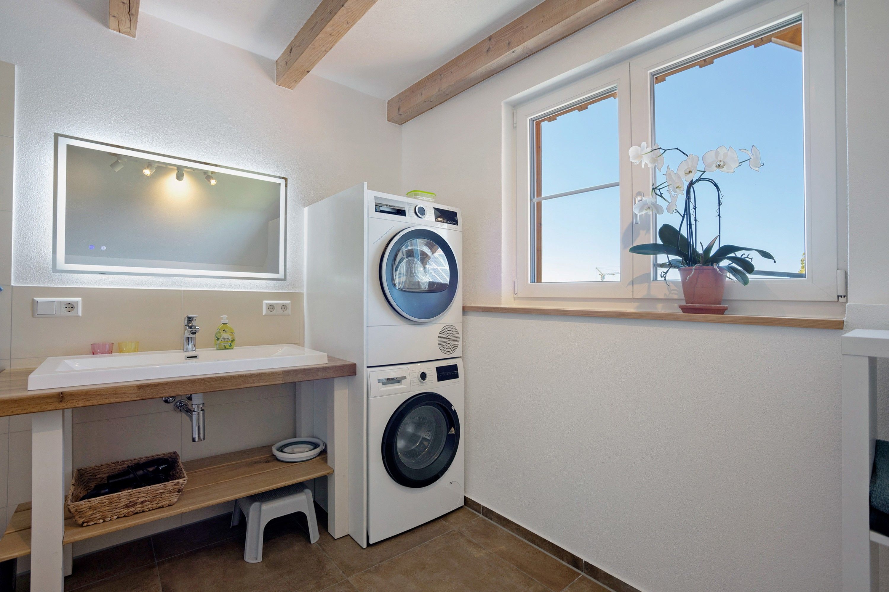 A laundry room with a washing machine, a sink, a mirror, and a window with an orchid plant.