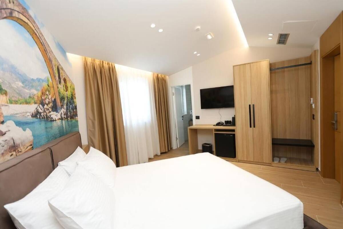 Ett hotellrum med en säng, en tv, ett skrivbord, en garderob och en tavla som föreställer ett landskap.