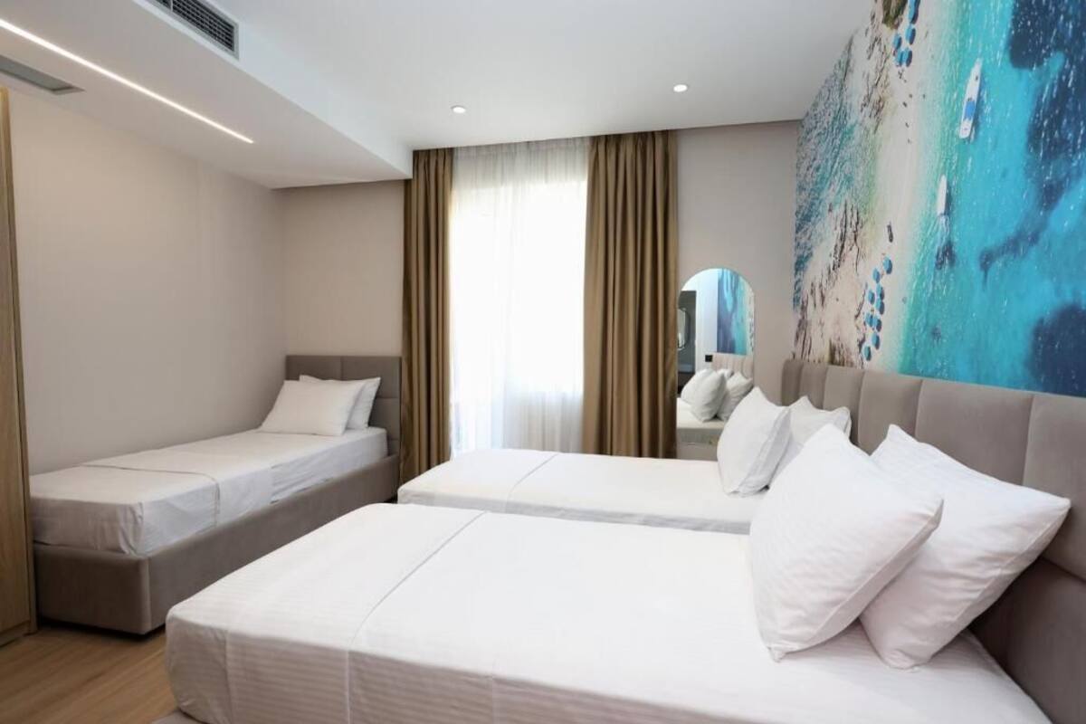 Ett hotellrum med två separata sängar, en stor säng, en tavla som föreställer en strandscen och ett fönster med gardiner.