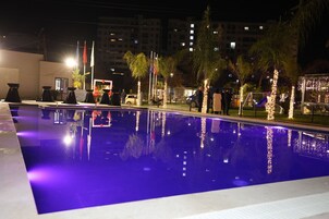 Μια πισίνα με κρυστάλλινα, γαλάζια νερά, περιτριγυρισμένη από φοίνικες και φωτισμένη με σειρές από φωτάκια.