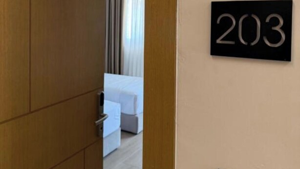 A hotel room with a wooden door, a number sign '203' on the wall, a bed, and a bedside table. - Vila Lira (Durres)