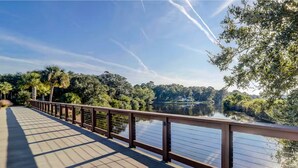 A wooden deck with a railing overlooking a calm lake surrounded by dense greenery. - Palmetto Dunes Beach Villa | Golf, Pool,  Dogs OK (Hilton Head Island)