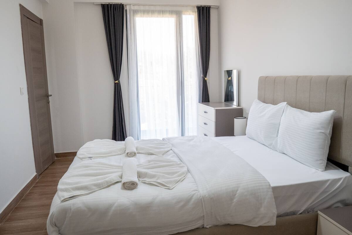 Een net opgemaakt bed met witte lakens, een nachtkastje met een lamp en een raam met gordijnen.