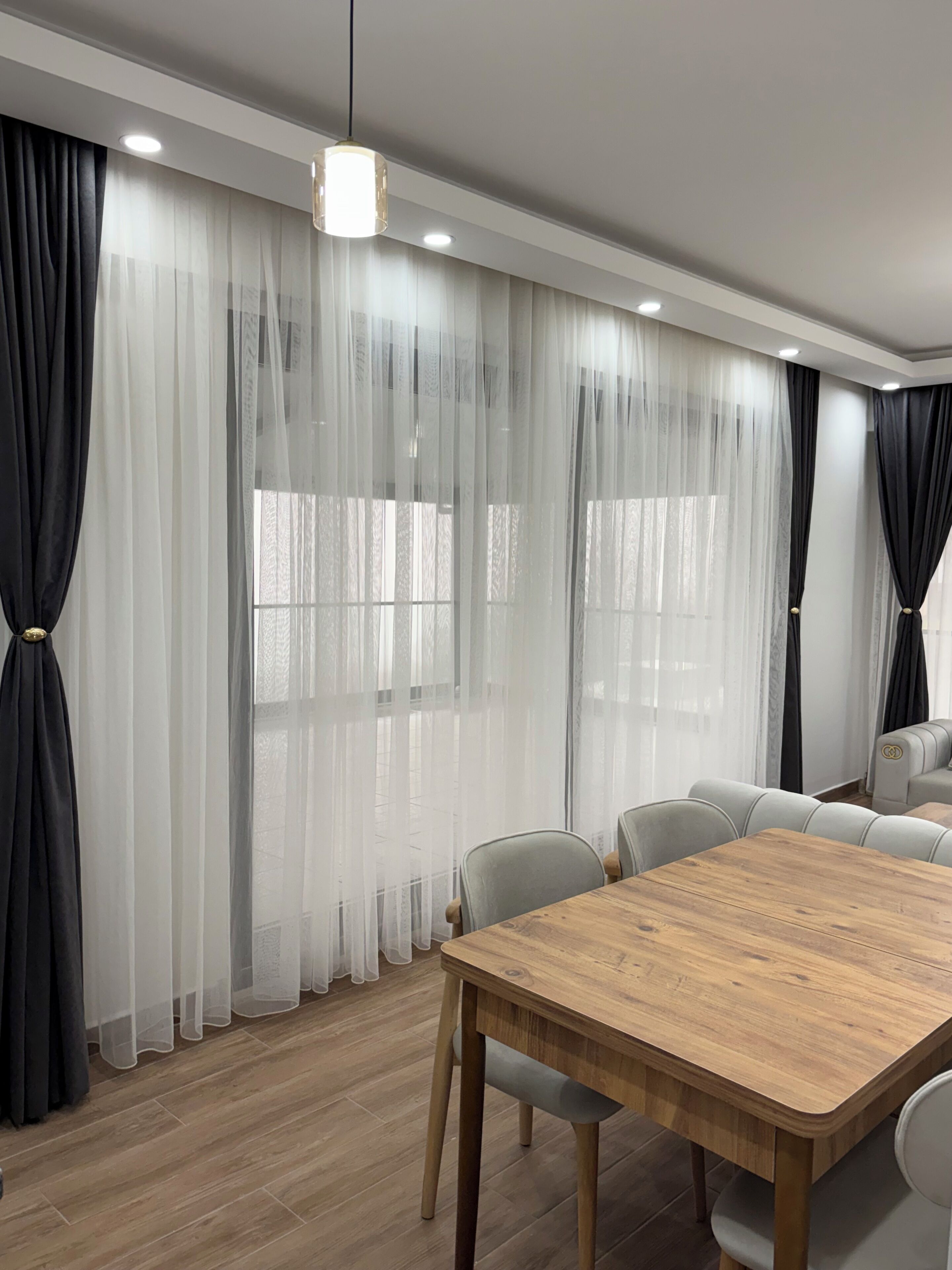 A modern dining area with a wooden table and chairs, sheer curtains, and a sofa.