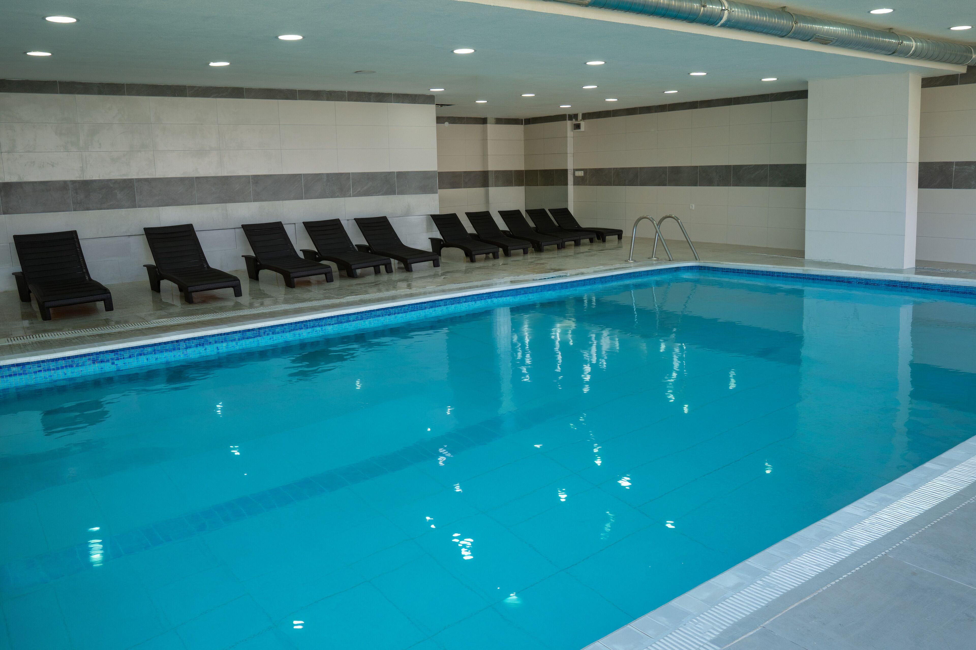 Ein Hallenbad mit klarem, blauem Wasser, schwarzen Liegestühlen und einer Wand mit grau-weißen Fliesen.