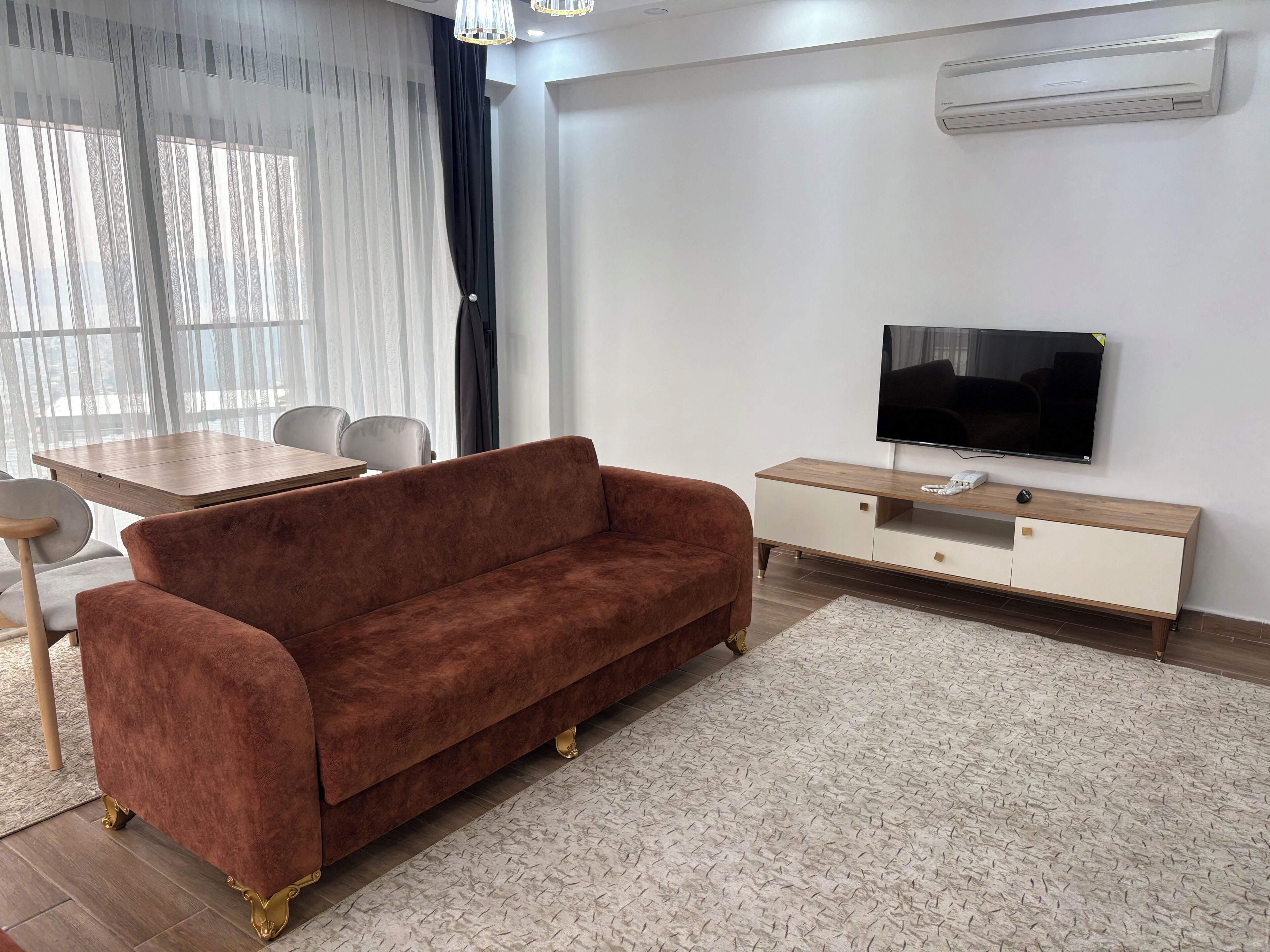 A living room with a brown sofa, a TV on a wooden stand, a dining table with chairs, and sheer curtains.
