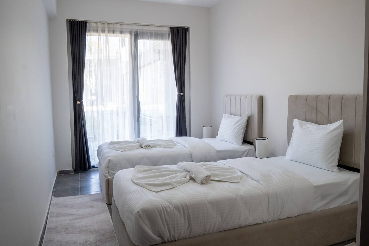 Een hotelkamer met twee bedden, een groot raam met dunne gordijnen, een nachtkastje met een lamp en een klein vloerkleed.