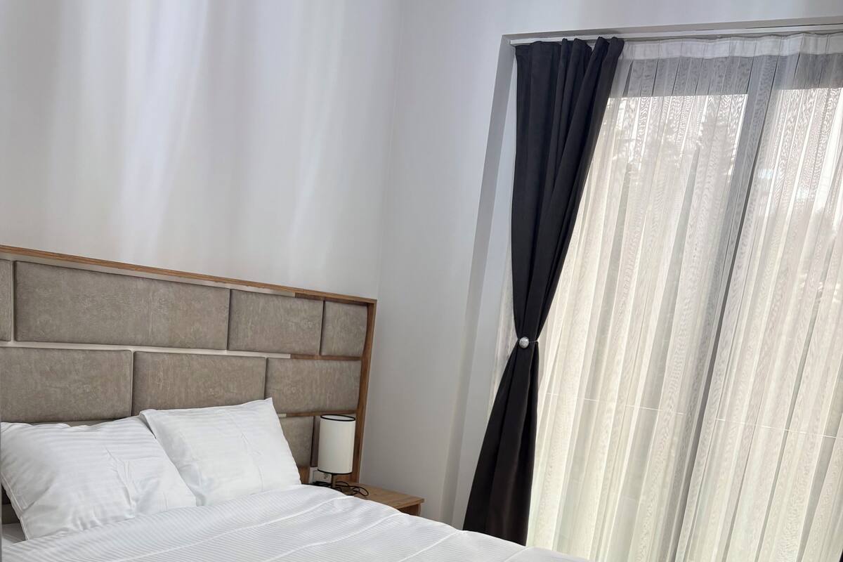 Een net opgemaakt bed met houten frame en witte beddengoed, een nachtkastje met een lamp, en een raam met dunne gordijnen en een donkere stof.