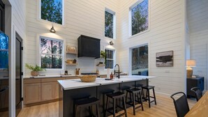 A modern kitchen with white wooden walls, a central island with bar stools, and large windows offering views of the surrounding forest. - Modern Chalet Retreat | 1BR + Loft & Bunks in Secluded Broken Bow (Broke Bow)