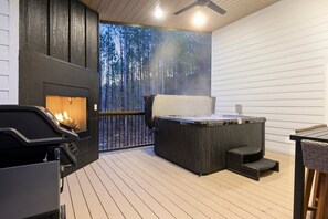 Um deck externo com uma lareira a lenha, uma banheira de hidromassagem e vista para uma floresta.