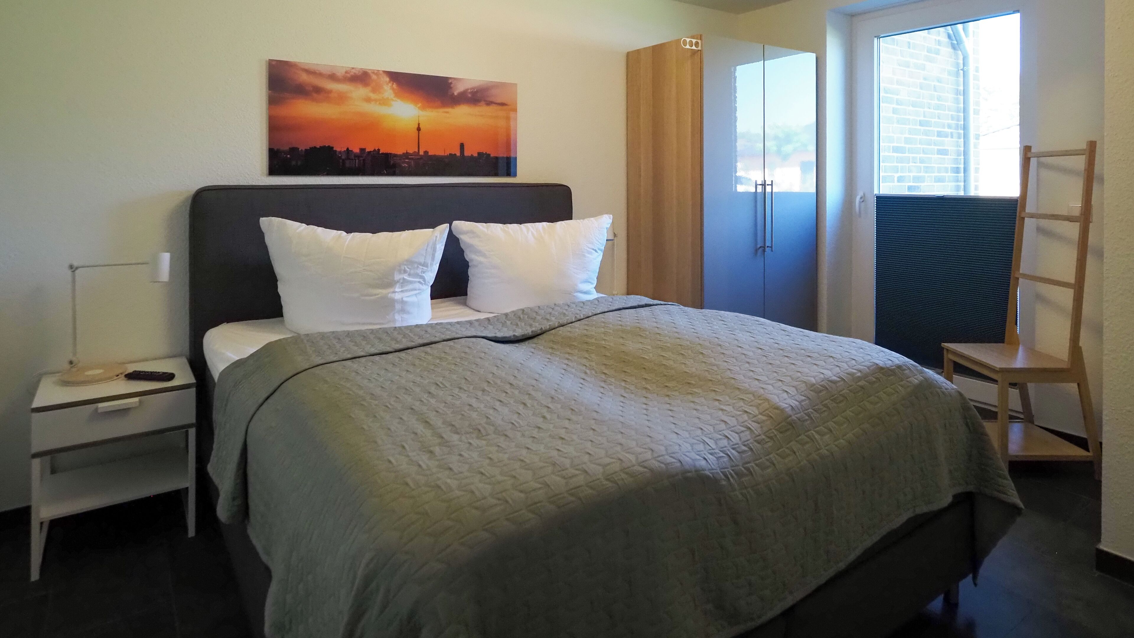 Une chambre d’hôtel avec un lit, une table de chevet, une lampe, un tableau représentant un coucher de soleil, une armoire et une fenêtre avec des stores.