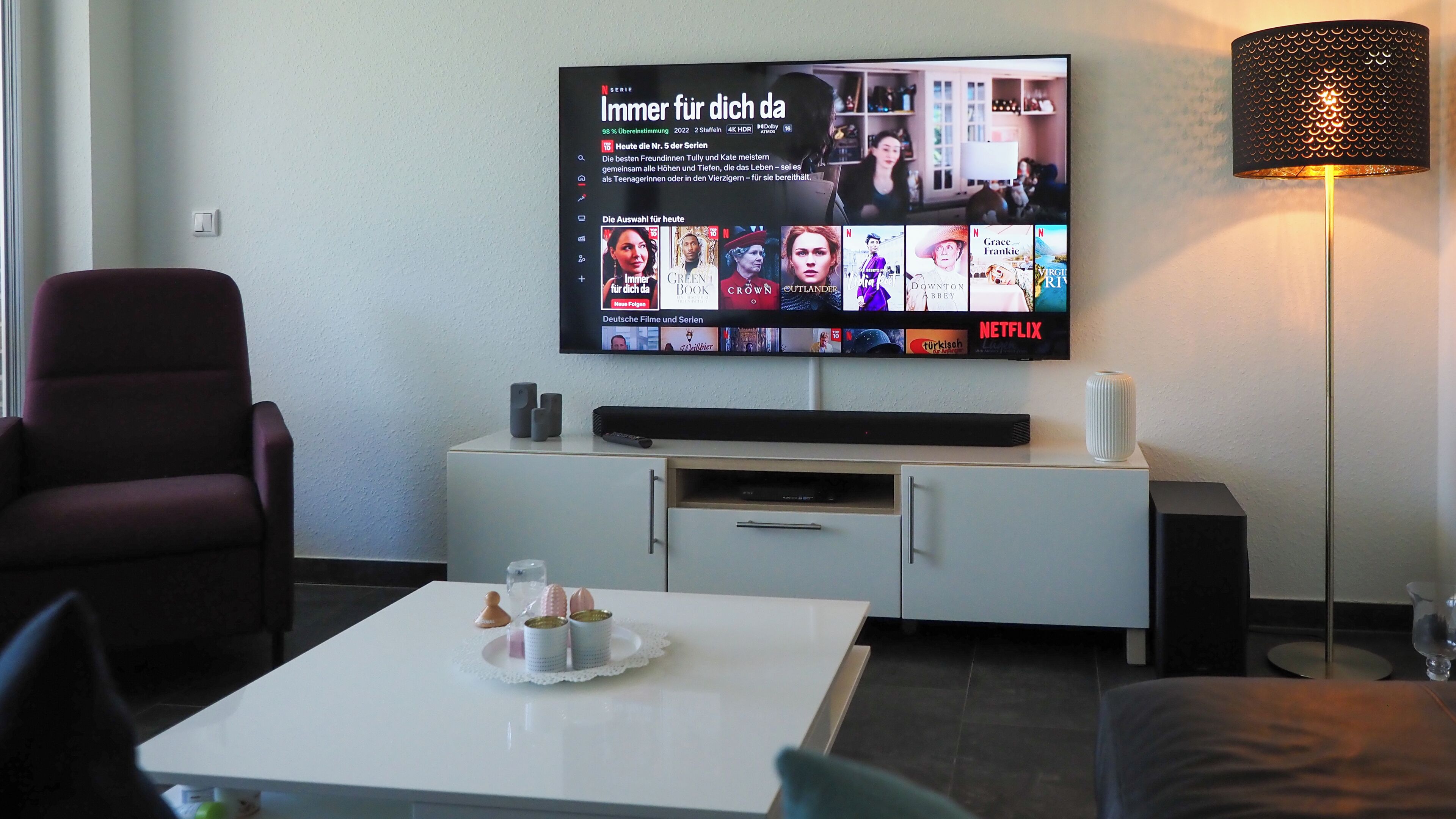 Ein modernes Wohnzimmer mit einem Flachbildfernseher, der eine Netflix-Oberfläche zeigt, einem weißen Couchtisch mit Dekorationen, einem roten Sessel und einer Stehlampe.