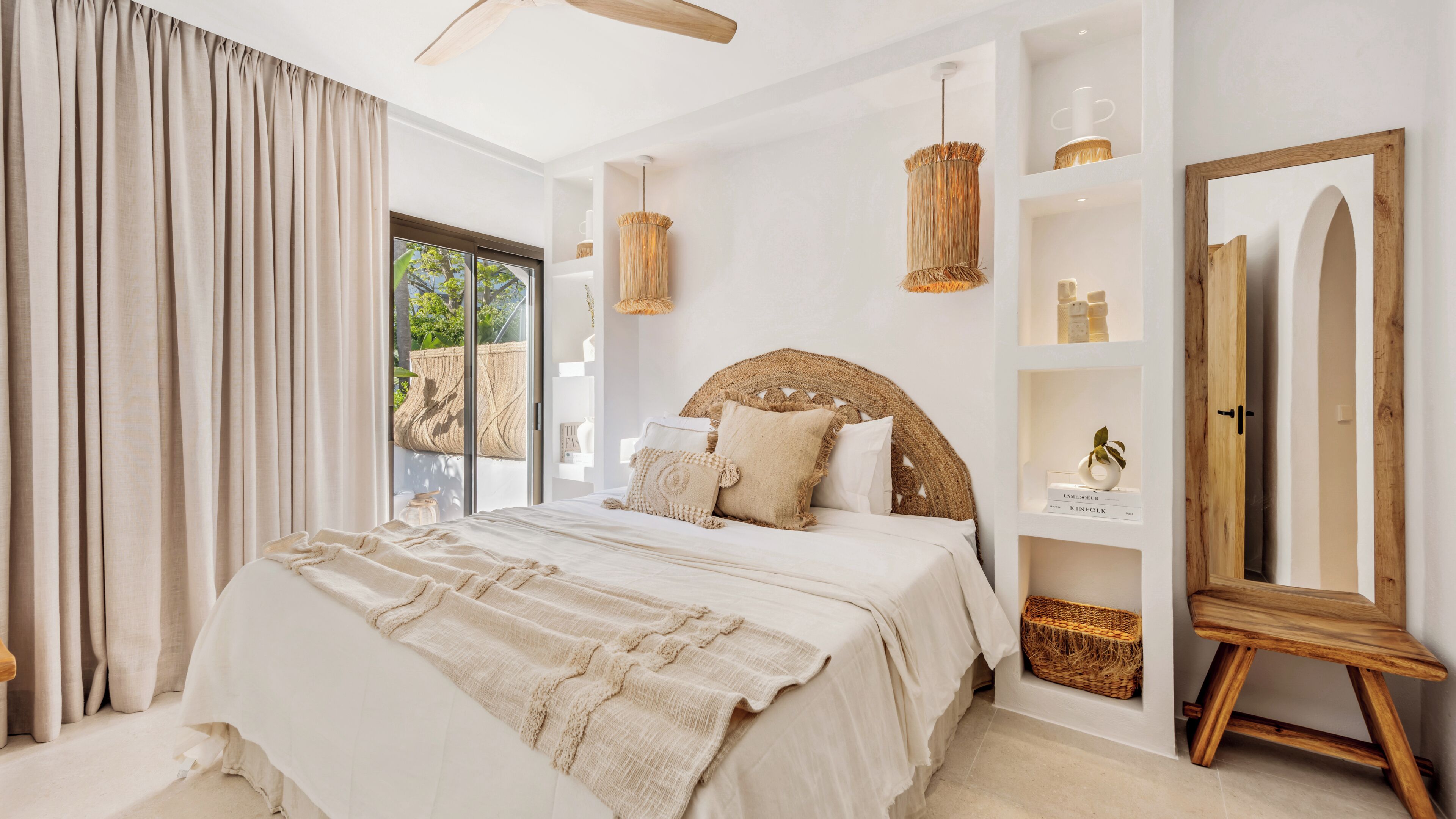 A bedroom with a large bed, a ceiling fan, a woven headboard, a bedside table with a lamp, and a view of greenery outside.
