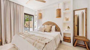 A bedroom with a large bed, a ceiling fan, a woven headboard, a bedside table with a lamp, and a view of greenery outside. - Beachside living in a charming urbanisation (Marbella)