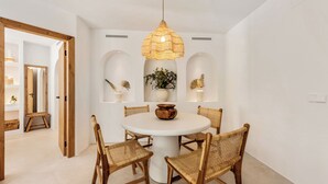 A dining area with a round table, wicker chairs, a hanging light fixture, and wall-mounted shelves with decorative items. - Beachside living in a charming urbanisation (Marbella)
