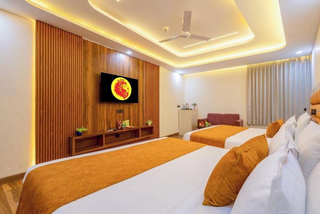 A hotel room with two beds, a TV mounted on a wooden panel wall, a ceiling fan, and a window with curtains.