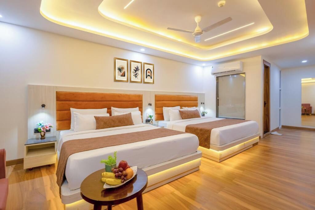A modern hotel room with two beds, a round table with fruit, a ceiling fan, and framed artwork on the wall.