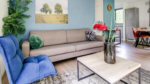 Un salón con sofá, una silla azul, una mesita de centro, un cuadro y una planta.