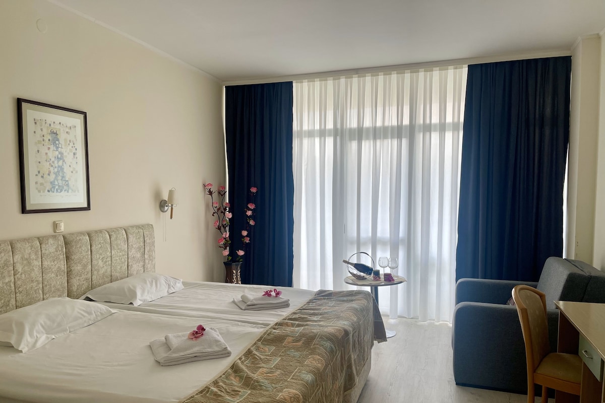 Habitación de hotel con cama, escritorio, silla, un florero con flores y un cuadro en la pared.