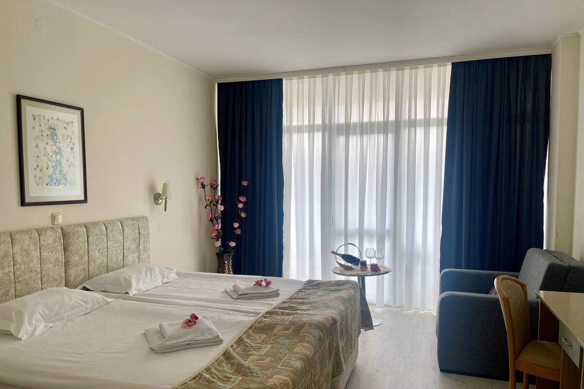 Habitación de hotel con cama, escritorio, silla, un florero con flores y un cuadro en la pared.