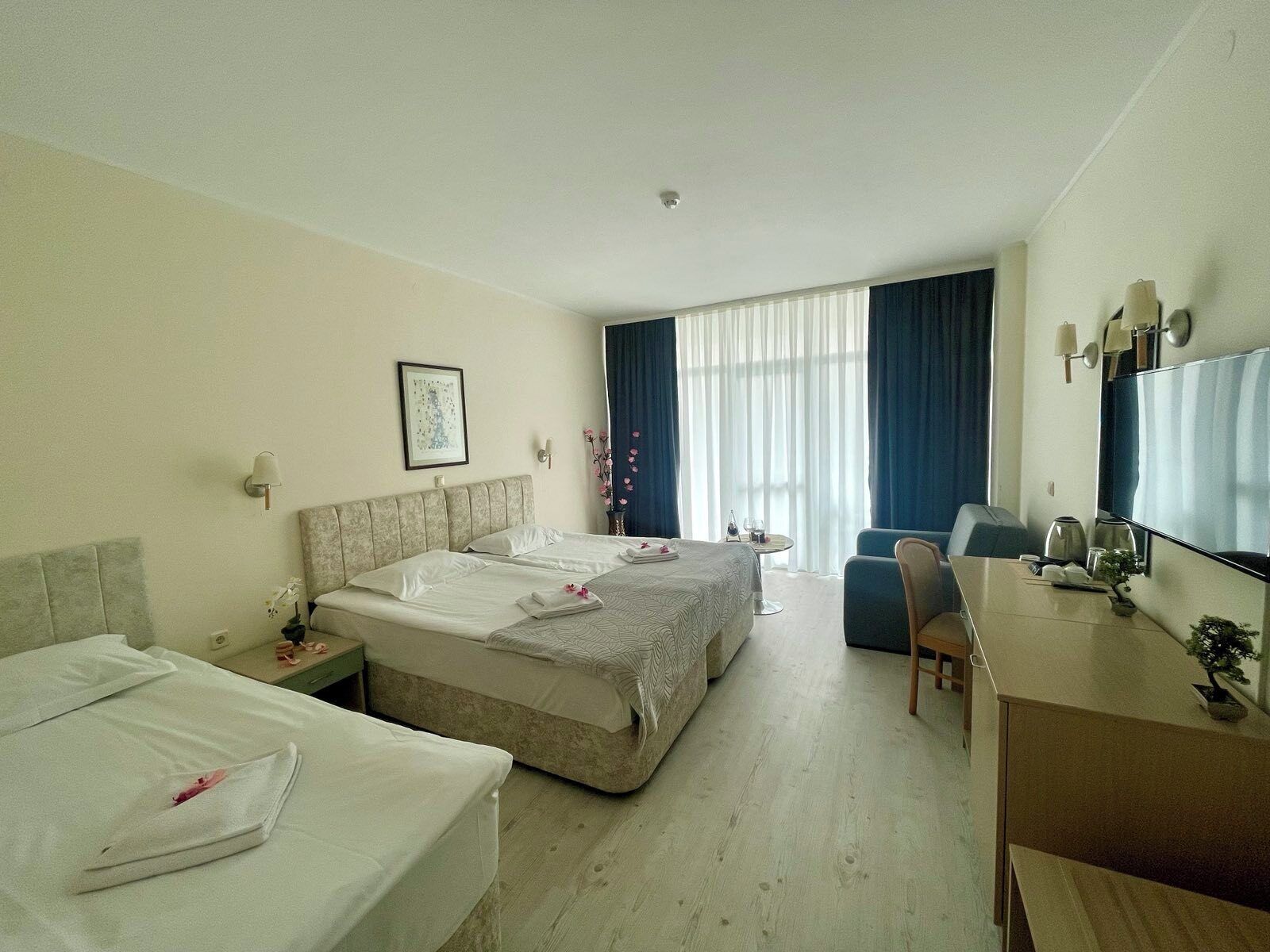 A hotel room with two beds, a desk, a chair, a small table with a wine glass, and a large window with curtains.