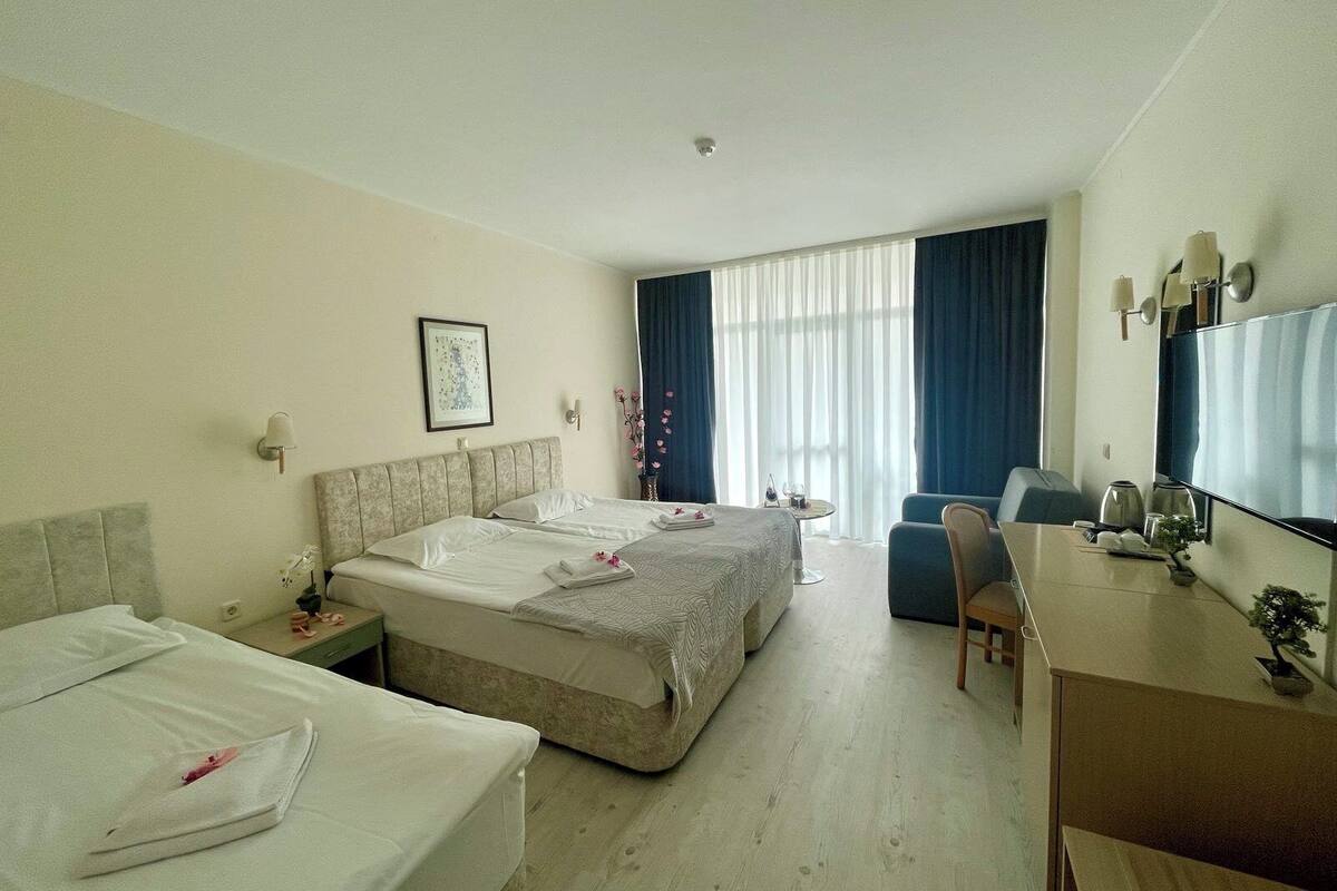 Habitación de hotel con dos camas, un escritorio, una silla, una mesita con una copa de vino y un gran ventanal con cortinas.